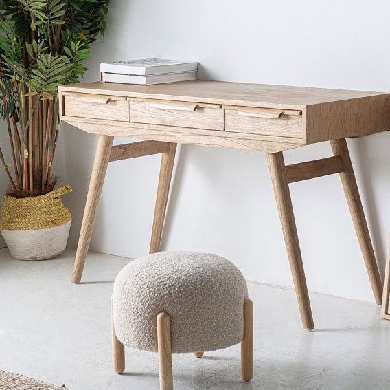 El Escritorio de madera mindi con tres cajones NYRY, de estilo nórdico, tiene patas anguladas; una planta está al lado y al frente hay un taburete redondo tapizado.