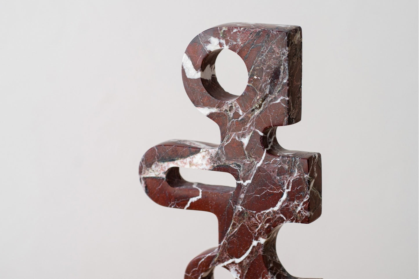 La Figura decorativa abstracta en mármol Stelio presenta una superficie roja oscura y brillante con vetas blancas, cortes circulares y rectangulares; Es perfecta como escultura moderna para decoración, exhibida sobre fondo liso.
