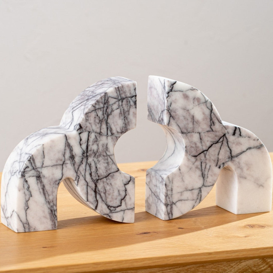Dos sujetalibros abstractos y curvos de Escultura de mármol blanco gris Aluna con vetas grises y negras descansan sobre una superficie de madera, añadiendo un toque escultórico moderno al fondo neutro.