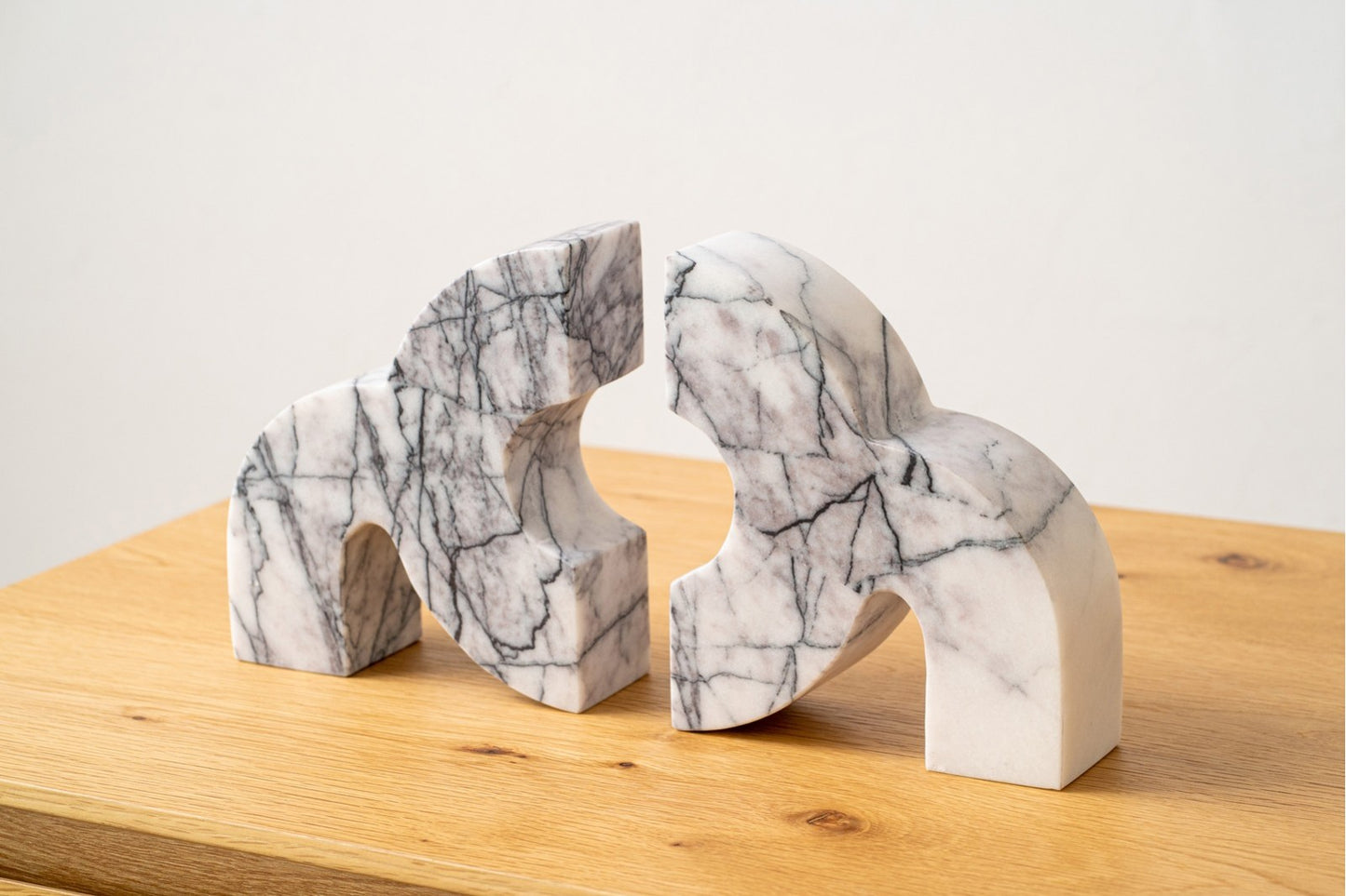 Dos esculturas de mármol blanco gris Aluna, con vetas negras y formas curvas y angulares, están dispuestas sobre una superficie de madera una frente a la otra formando una llamativa figura decorativa.