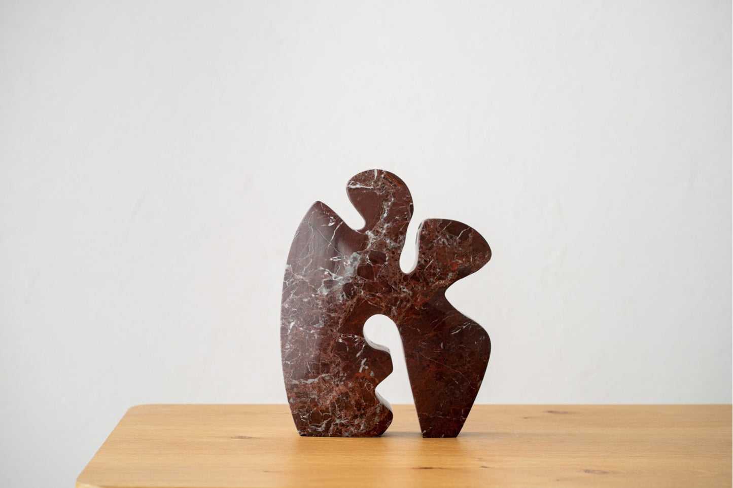 La Escultura en mármol rojo Mar N1 presenta suaves curvas y recortes en mármol rojo oscuro, que descansan sobre una superficie de madera clara para crear una llamativa pieza moderna sobre un fondo blanco liso.
