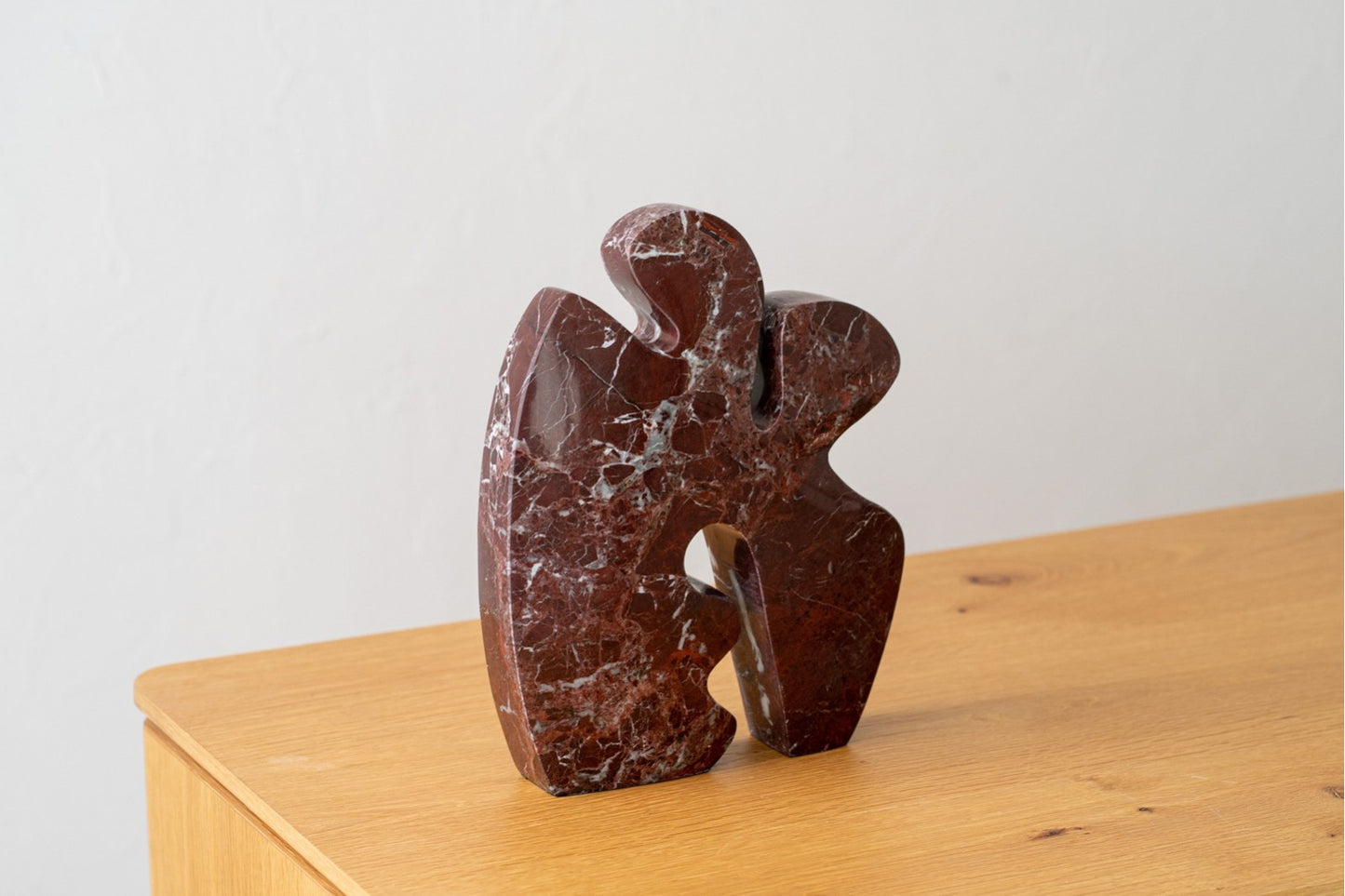 La Escultura en mármol rojo Mar N1 presenta formas abstractas curvas y recortadas en mármol rojo oscuro, colocadas sobre una superficie de madera clara con un fondo blanco: una sorprendente figura decorativa y escultura moderna para espacios contemporáneos.
