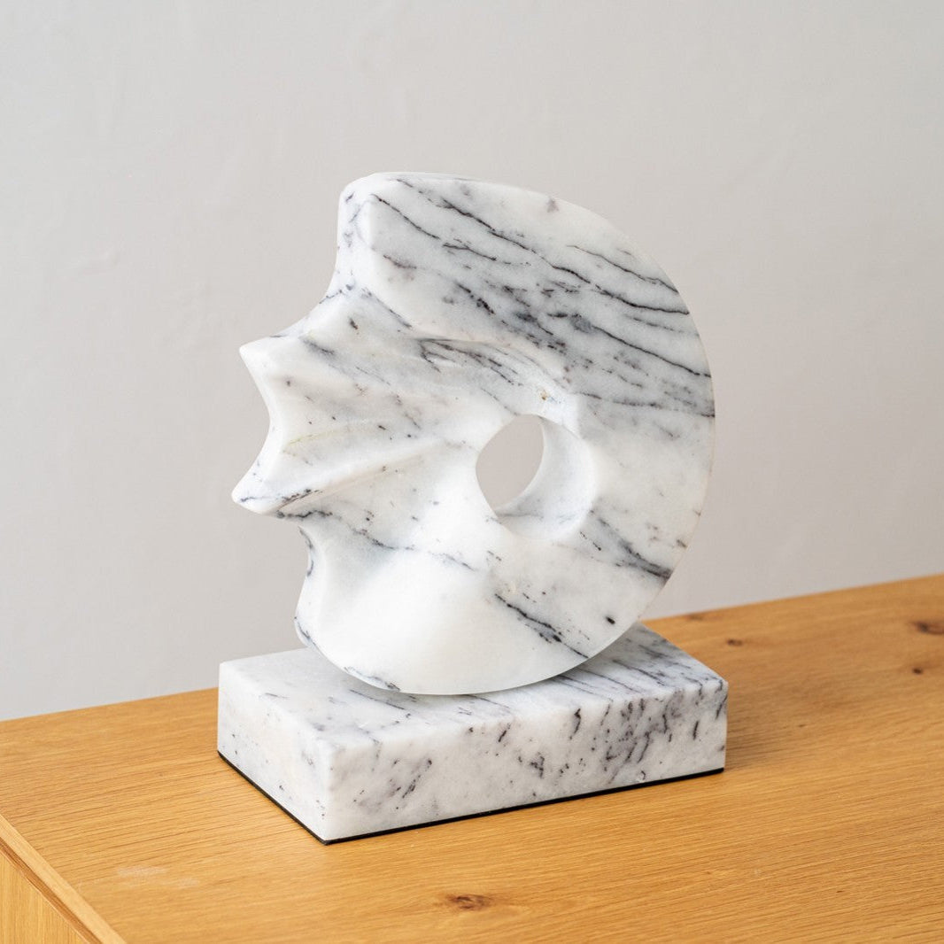 La Escultura en mármol Mar N2 destaca por su elegancia natural, vetas negras y diseño circular con borde irregular y agujero central, montada sobre base rectangular. Perfecta para decorar interiores y realzar superficies de madera.