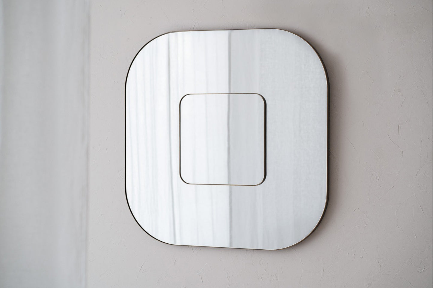 Un moderno espejo de pared cuadrado doble de metal 89×4×89 CM en oro envejecido con diseño de doble marco refleja suavemente cortinas, montado sobre una pared texturizada clara.