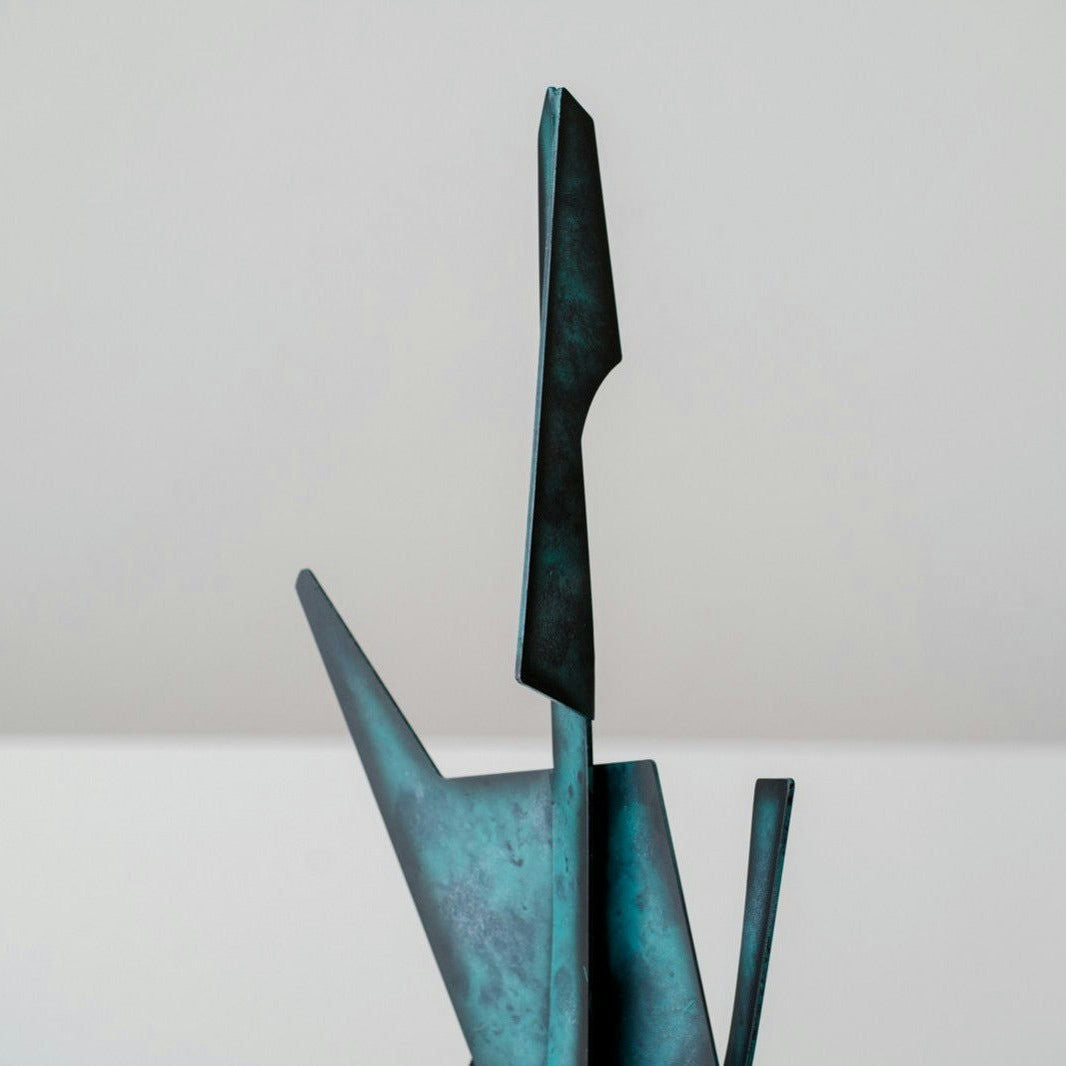 La Figura Decorativa en Metal Mármol Abstracción N2 es una escultura abstracta de metal con formas angulares y alargadas y una pátina azul verdosa, que agrega elegancia sofisticada a cualquier espacio cuando se coloca sobre un fondo liso.