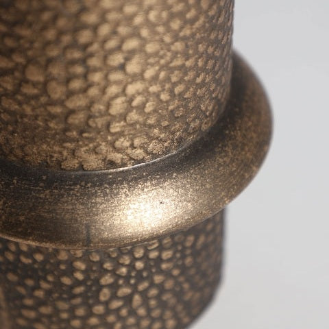 Primer plano de un recipiente cilíndrico Vesc E texturizado de color bronce con un diseño art déco, que presenta una banda brillante y exuda un refinado arte decorativo.