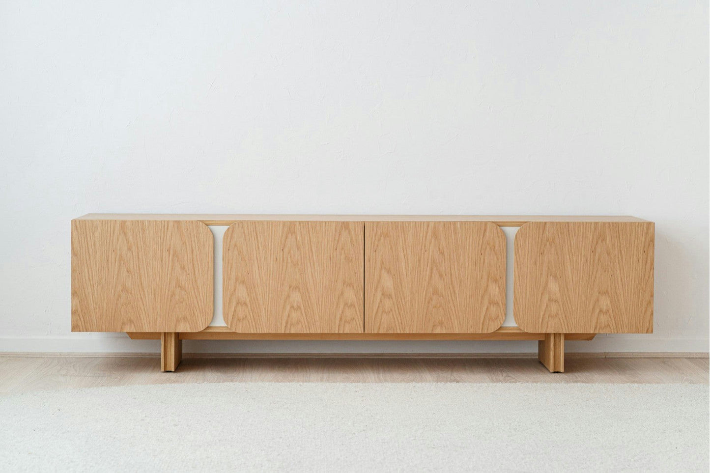 Un aparador minimalista de madera alargado con puertas de esquinas redondeadas, con un acabado en madera clara y acabado arena mate, se encuentra sobre un piso claro contra una pared blanca lisa. Similar al Mueble TV en DM y roble natural detalles gris arena Curva.