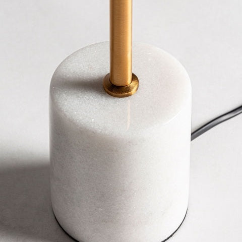 Primer plano de la Lámpara de sobremesa en mármol y metal Leba W, que presenta una base lisa de mármol blanco, una estructura de varillas de metal dorado en el centro y un cable eléctrico negro visible en el lateral.