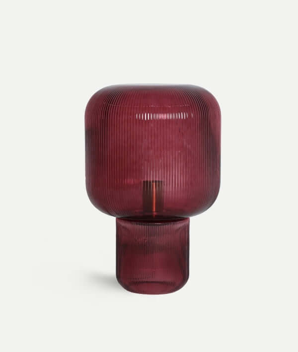 La Lámpara de sobremesa cristal burdeos estriado 27×27×42 cm - moderna - SIRA B presenta una pantalla de cristal burdeos estriado y una base cilíndrica, sobre un fondo blanco liso.