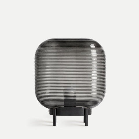 La lámpara de mesa en vidrio ahumado Cielo destaca por su elegante diseño, con pantalla redondeada de vidrio negro ahumado y base corta de metal negro, ideal para ambientes con fondo claro y sencillo.