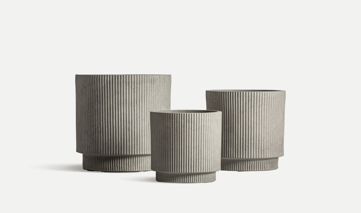 Tres maceteros de cerámica estriada gris de diferentes tamaños del Juego VULK se exhiben sobre fondo blanco, ideales para interiorismo y aportar un toque contemporáneo y elegante a cualquier espacio.