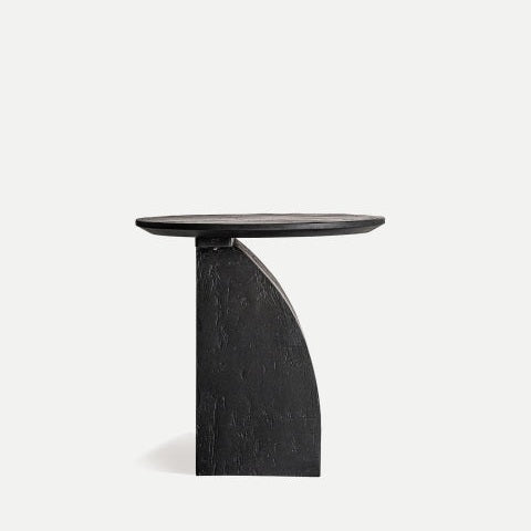 La mesa auxiliar de madera de mango Annika es una moderna mesa redonda negra con base geométrica asimétrica, elaborada con madera de mango resistente. Se destaca elegantemente sobre un fondo claro.