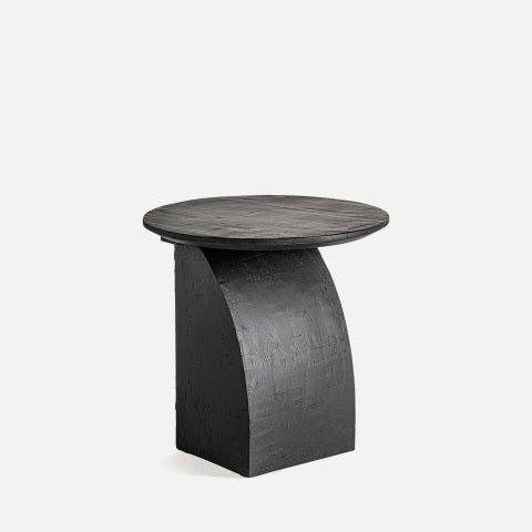 La mesa auxiliar de madera de mango Annika presenta un diseño moderno en color negro con una parte superior redonda y una base rectangular curva, sobre un fondo gris claro.