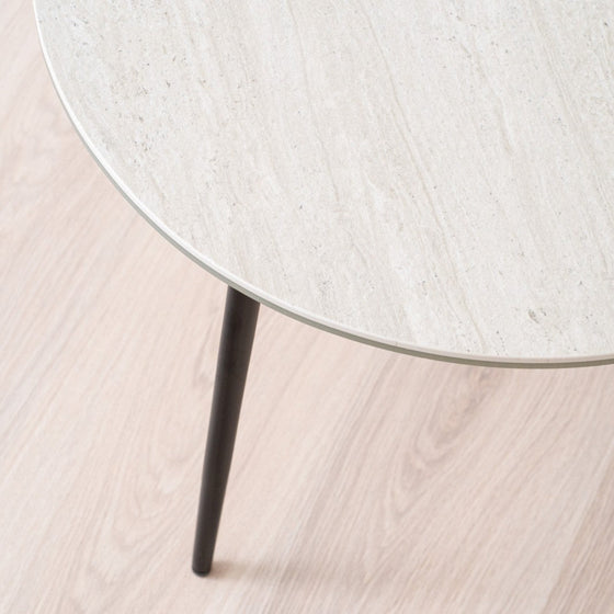 La Mesa auxiliar Redonda en Cerámico gris Erma Ø60, con superficie de madera clara y una única pata de metal negro, complementa el suelo de madera a juego, derrochando diseño cómodo.