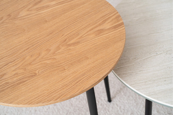 La mesa auxiliar Redonda en Cerámico gris Erma Ø60 presenta una elegante textura de piedra gris y patas de metal negro, junto con una mesa con un acabado de veta de madera clara. Ambas exudan un diseño sofisticado sobre una alfombra clara, agregando elegancia a cualquier espacio.