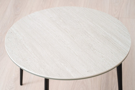 La Mesa auxiliar Redonda en Cerámico gris Erma Ø60 presenta una superficie beige claro con patas negras sobre un piso de madera clara, creando un aspecto sofisticado perfectamente complementado con cerámica gris mate para mayor elegancia.