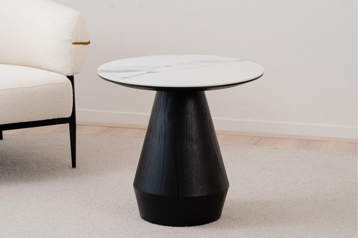 Una mesa auxiliar redonda contemporánea, la FRES 60×55 CM, presenta un diseño de cerámica y madera de fresno y se alza sobre una alfombra clara junto a una silla tapizada en color crema.