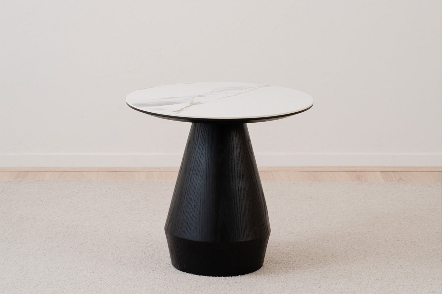 La Mesa auxiliar redonda de cerámica y madera de fresno 60×55 CM FRES, de estilo contemporáneo, se exhibe sobre una alfombra clara en una habitación minimalista.