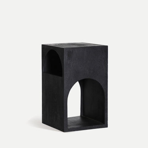 La mesa auxiliar cuadrada en madera de mango Kirila es un taburete geométrico negro con dos aberturas arqueadas en cada lado, que presenta un diseño minimalista y está elaborado con madera de mango duradera.