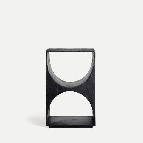 La mesa auxiliar de madera de mango Kirila, elaborada con madera de mango negro, presenta cortes semicirculares opuestos que forman una elegante forma de reloj de arena. Sobre un fondo blanco roto, ejemplifica el diseño contemporáneo.