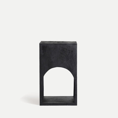 La mesa auxiliar cuadrada en madera de mango Kirila es un bloque de madera negro con forma rectangular y un recorte arqueado cerca de la base, elaborado en un estilo de diseño minimalista, sobre un fondo claro y liso.