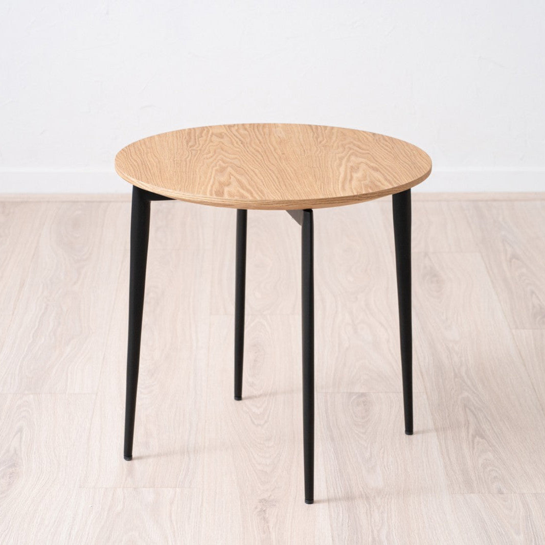 La Mesa auxiliar Redonda en fresno natural y Metal Edgar Ø50 cuenta con un diseño de madera fina con elegantes patas de hierro negro, que complementan el piso natural y el fondo blanco.