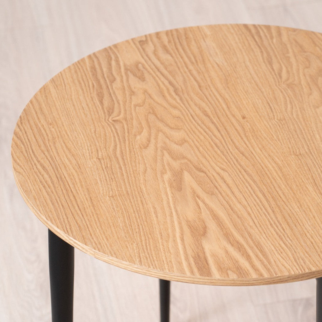 La Mesa auxiliar Redonda en fresno natural y Metal Edgar Ø50 presenta un patrón de veta natural de fresno natural con elegantes patas de hierro negro, perfectas en un piso de madera clara.