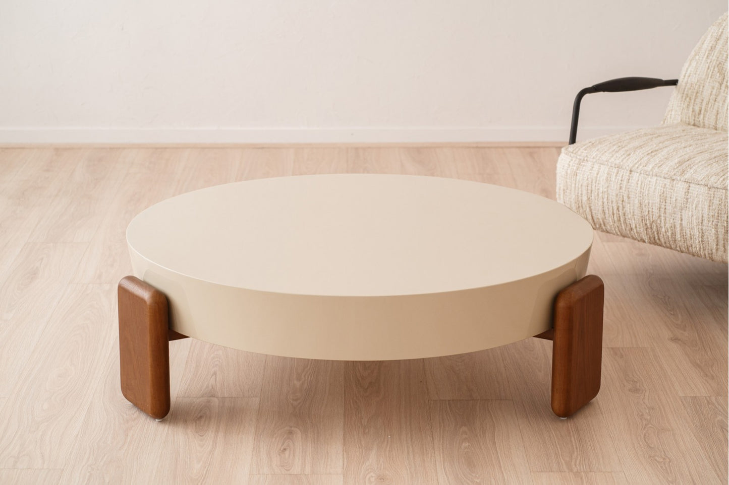 A Mesa de centro redonda de madera de nogal lacado beige 110×110×35 cm OBI descansa sobre un piso de madera clara junto a una silla beige tapizada con reposabrazos negros.