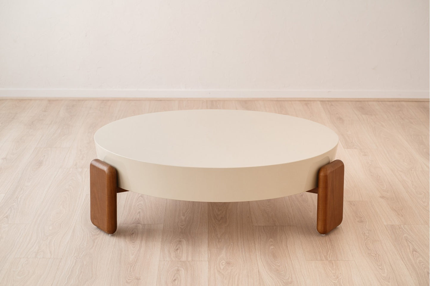 A Mesa de centro redonda de madera de nogal - lacado beige 110×110×35 CM - moderno - OBI con tres robustas patas de nogal se alza sobre un suelo de madera clara junto a una pared blanca lisa.