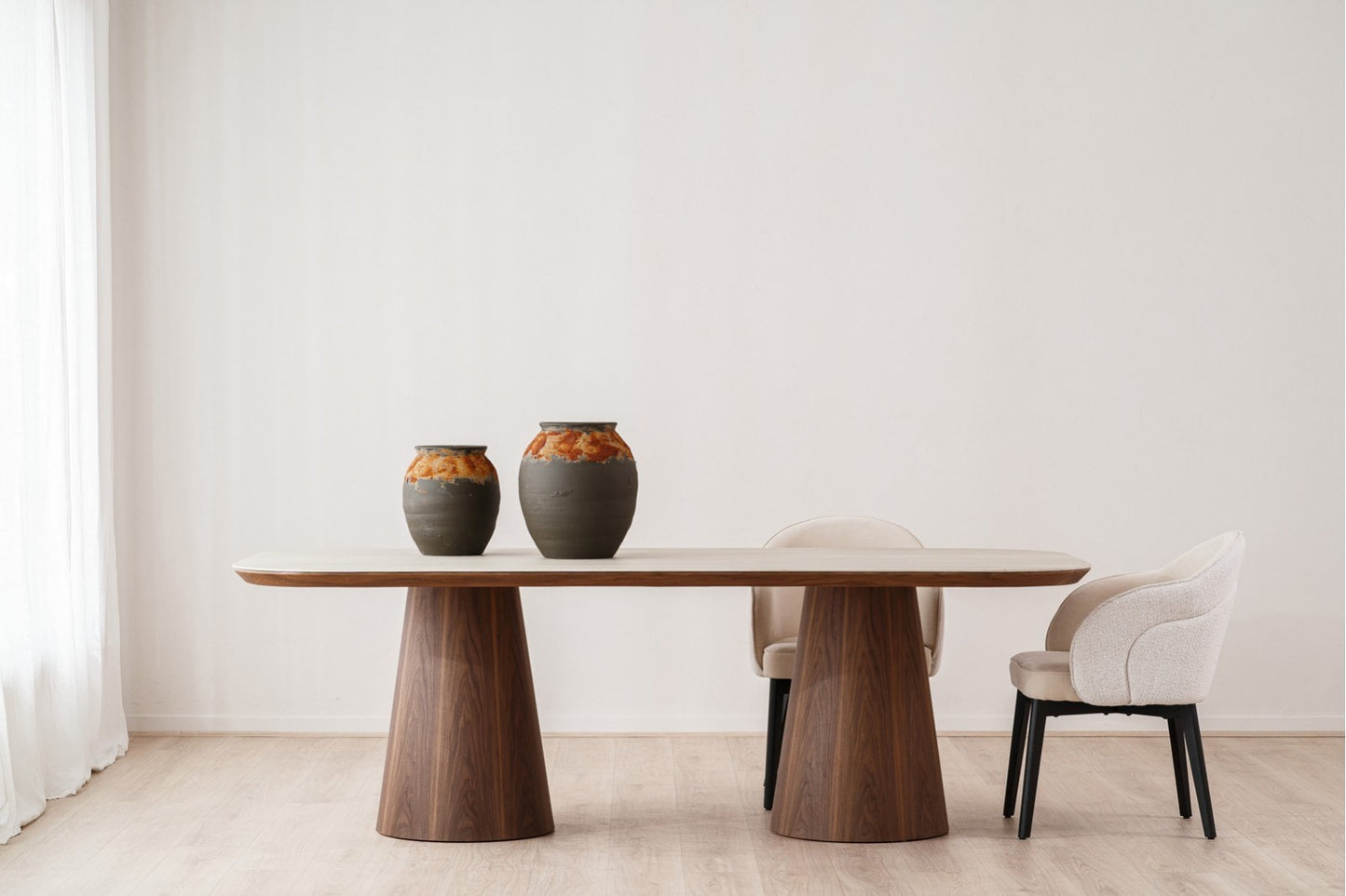 Un comedor minimalista muestra la mesa de comedor de cerámica y madera de nogal DOLMEN (230×110×76 CM) con dos sillas beige y dos jarrones de cerámica, todo ello sobre una pared blanca lisa.