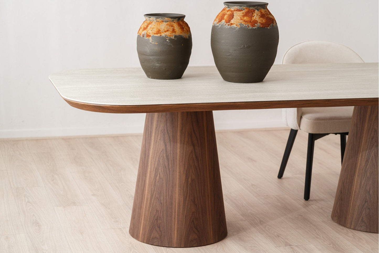 A mesa contemporánea de comedor de cerámica y madera de nogal (230×110×76 cm, modelo DOLMEN), con dos jarrones cerámicos y una silla tapizada beige sobre suelo de madera clara.
