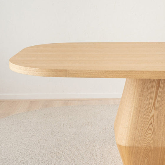 La gran mesa de comedor Evita (230 x 117 cm) con esquinas redondeadas descansa delicadamente sobre una alfombra texturizada y clara. Su base geométrica y gruesa de fresno natural encarna un diseño versátil que se integra perfectamente en cualquier ambiente con un atractivo fresco.