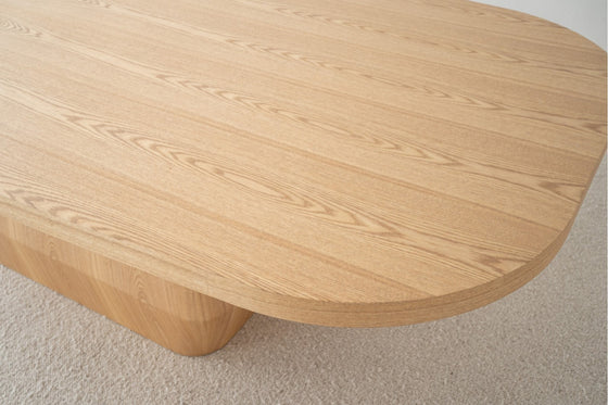 Primer plano de una mesa de madera con bordes redondeados sobre un suelo de moqueta clara. La mesa comedor (230x117cm) en DM y fresno natural Evita, parte de la colección Fresco Natural, presenta un patrón de vetas de madera natural que complementa su diseño versátil.