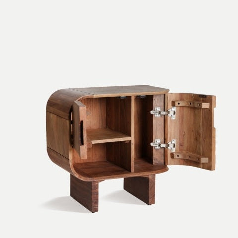 La mesita de noche Blesle de madera de acacia presenta una puerta abierta que revela estantes interiores y combina un diseño redondeado con bisagras metálicas. Fabricada en madera de acacia, ofrece un almacenamiento práctico y añade elegancia a cualquier habitación.