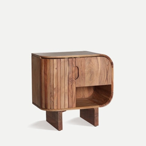 La Mesita de noche en madera de acacia Blesle ofrece un diseño contemporáneo con una estructura de bordes curvados, una puerta de gabinete y un estante abierto sobre dos patas rectangulares.