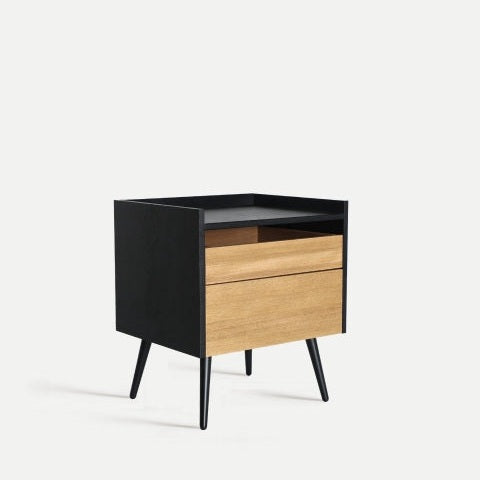 La mesita de noche en madera MDF y hierro Brussa es una elegante mesita de noche con un diseño contemporáneo, que cuenta con dos cajones de madera y un marco y patas negros, bellamente colocados sobre un fondo claro y liso.