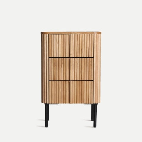La mesita de noche en madera y hierro Skagen, elaborada con madera de paulownia duradera con ranuras verticales y tres cajones, descansa sobre cuatro patas de metal negro, mostrando un estilo nórdico limpio sobre un fondo claro y liso.