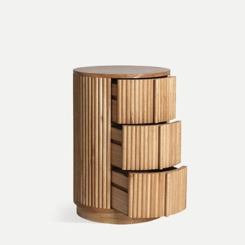 La mesita de noche en madera de paulownia Skagen2 es una mesa auxiliar cilíndrica de madera con ranuras verticales y tres cajones parcialmente abiertos, que ejemplifica un diseño nórdico minimalista que combina a la perfección forma y función.