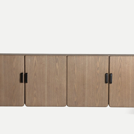 El mueble de TV en madera de fresno Busk es un mueble de madera largo y elegante con cuatro puertas cerradas y tiradores negros sobre un fondo blanco liso, que muestra un diseño moderno con almacenamiento integrado.