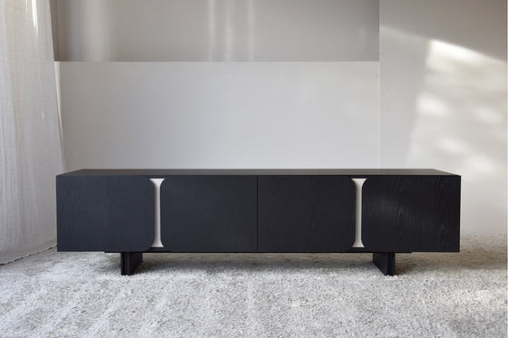 En una habitación minimalista, el Mueble TV Roble Negro mate Y Arena - Curva con su elegante diseño de curva y patrones geométricos se destaca sobre una alfombra clara, ejemplificando el diseño contemporáneo.