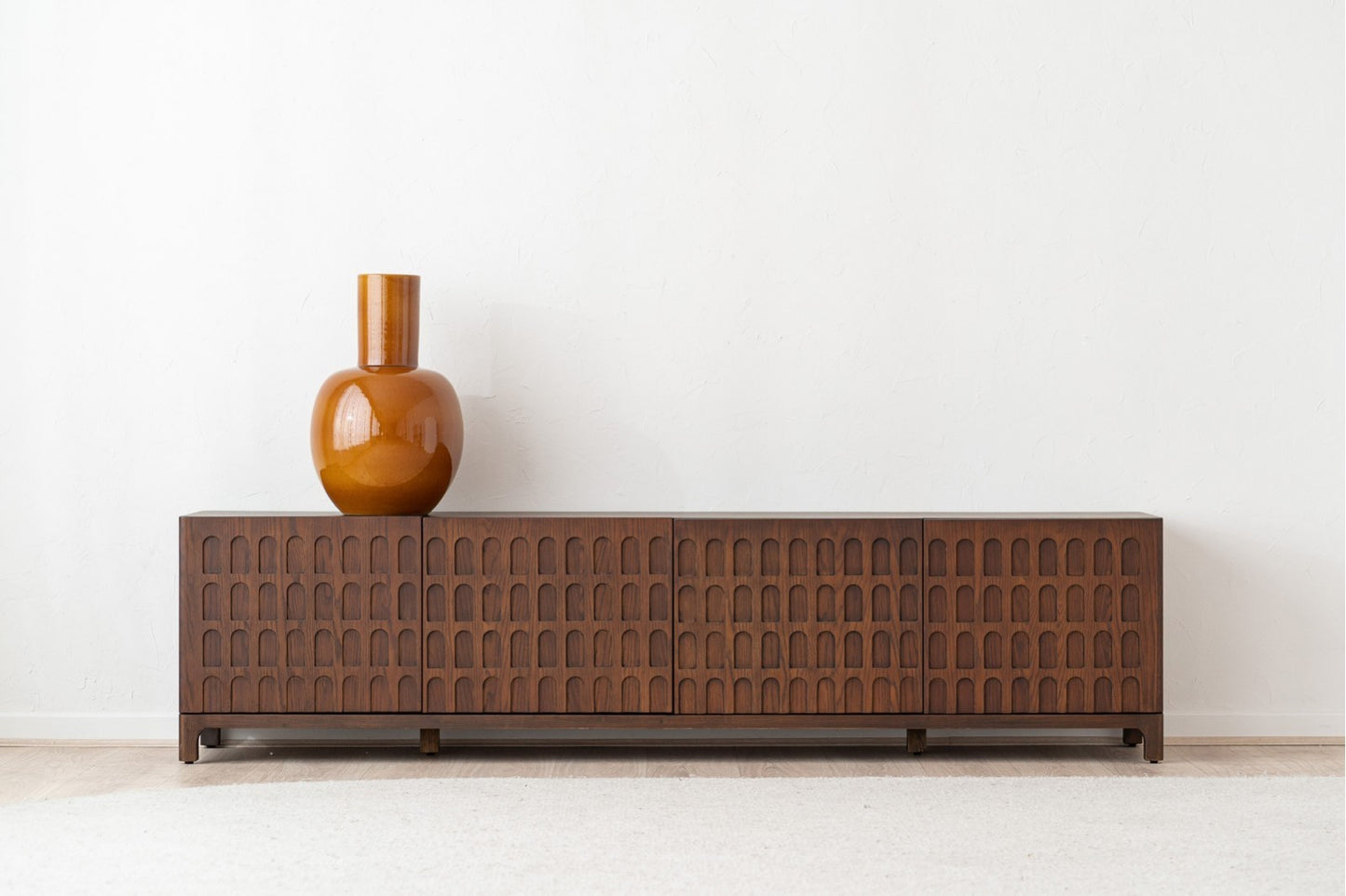 Sobre el Mueble TV de madera de roble oscuro 210×40×52 CM - contemporáneo - DARA, con un diseño de arcos que se repite, hay un gran jarrón de color ámbar colocado contra una pared blanca lisa.
