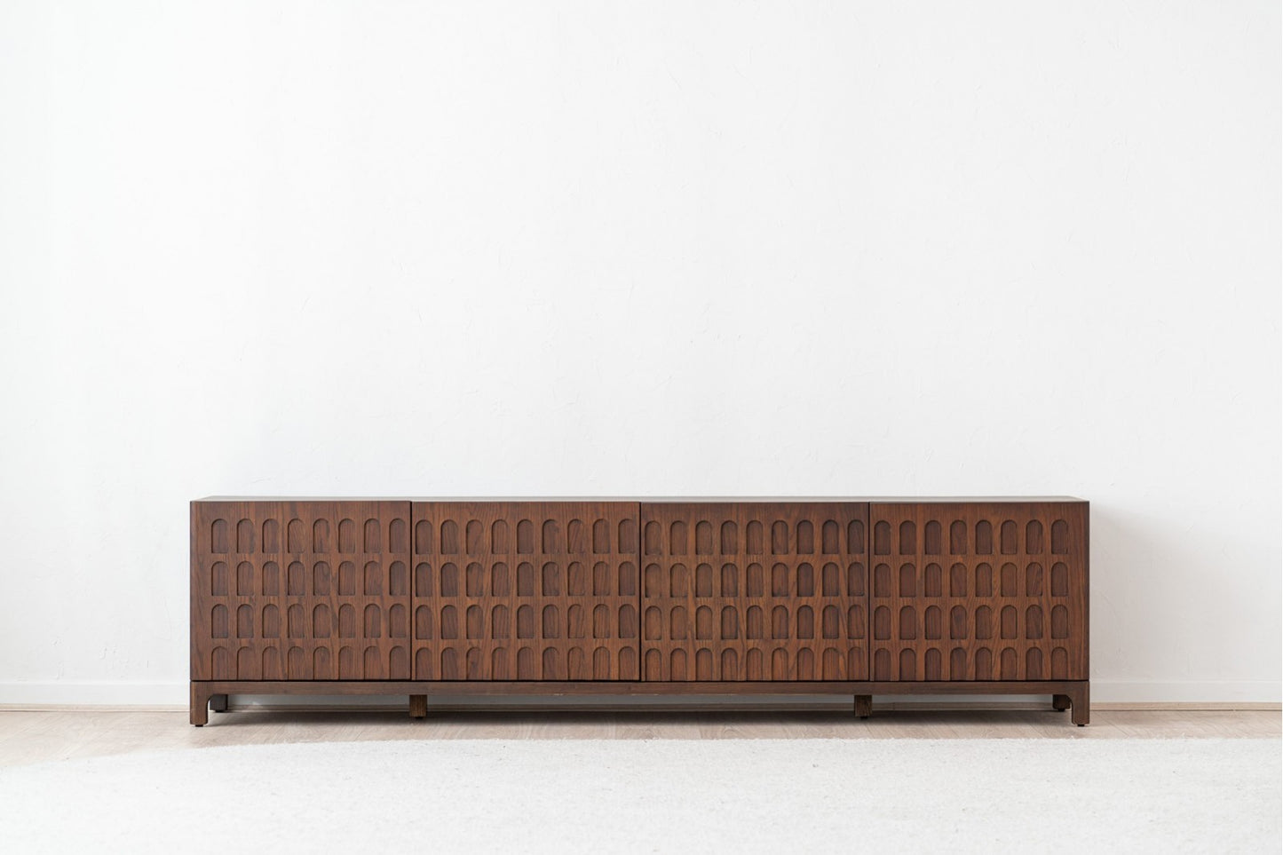 Mueble TV de madera de roble oscuro DARA (210×40×52 cm), bajo y contemporáneo, con arcos repetidos en las puertas, sobre una pared blanca lisa y un suelo claro.