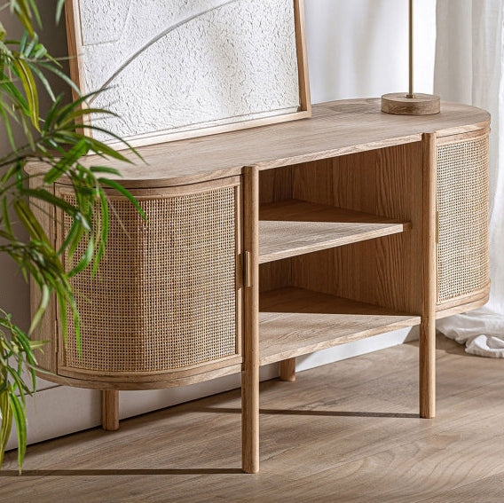 Un aparador Ikla de Mueble TV en DM con fresno cuenta con estantes abiertos y elegantes puertas de caña, realzadas por un panel decorativo con textura en la parte superior. Esta impecable combinación de materiales queda perfecta junto a una planta exuberante.
