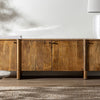 El Mueble TV en madera de mango Koigi es un aparador de madera con un acabado liso, elaborado con madera de mango resistente. Cuenta con dos gabinetes centrales, elegantes tiradores negros y exhibe un estilo nórdico que contrasta con elegancia sobre una pared blanca.