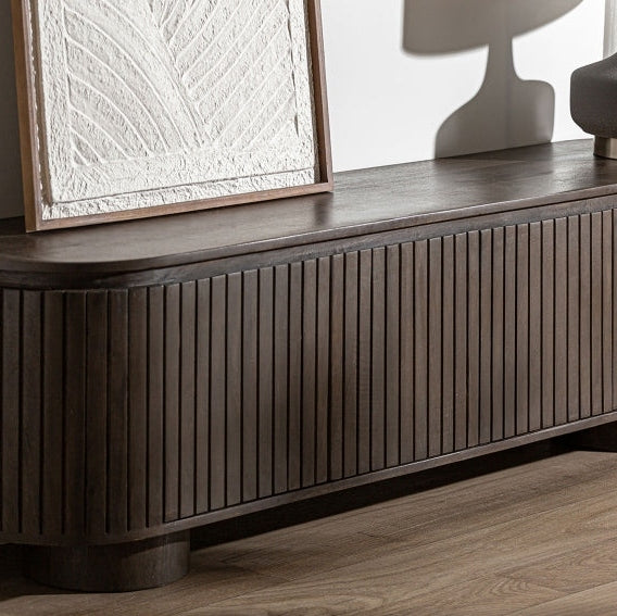 El Mueble para TV en madera de mango Kolga, un mueble oscuro con ranuras verticales elaborado en exquisita madera de mango, reposa contra la pared. Una obra de arte texturizada enmarcada y una lámpara de mesa descansan sobre la pared, integrándose a la perfección con el diseño contemporáneo de la habitación.