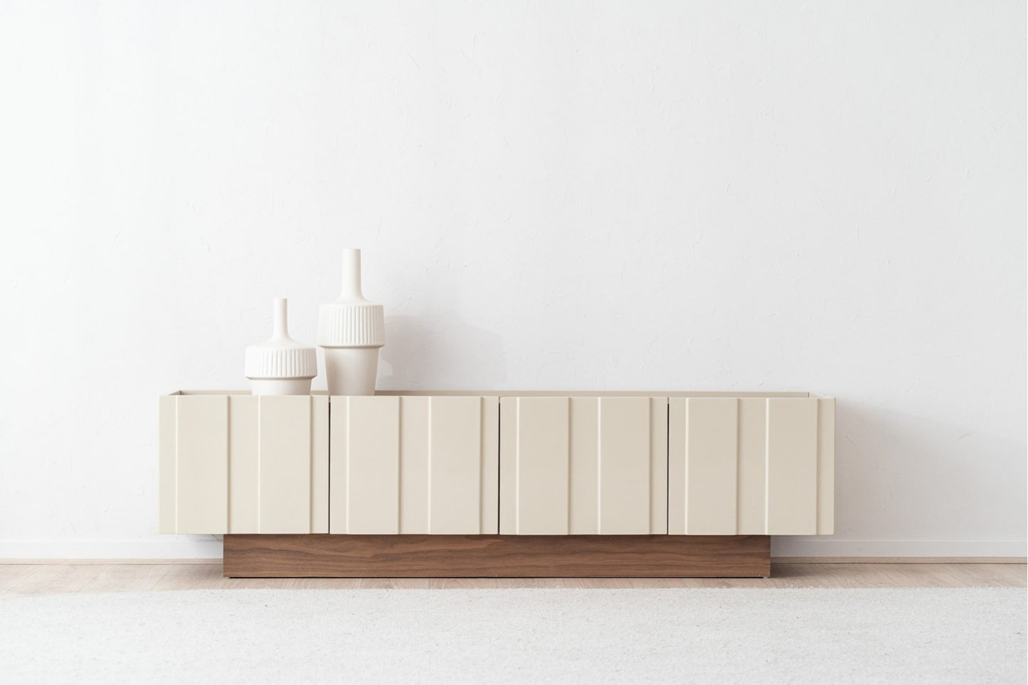 A moderno mueble TV de madera de nogal lacado beige brillo 198×45×53 cm OBI con detalles minimalistas y ranuras verticales se apoya en una pared blanca, decorado con tres jarrones blancos de diferentes alturas.
