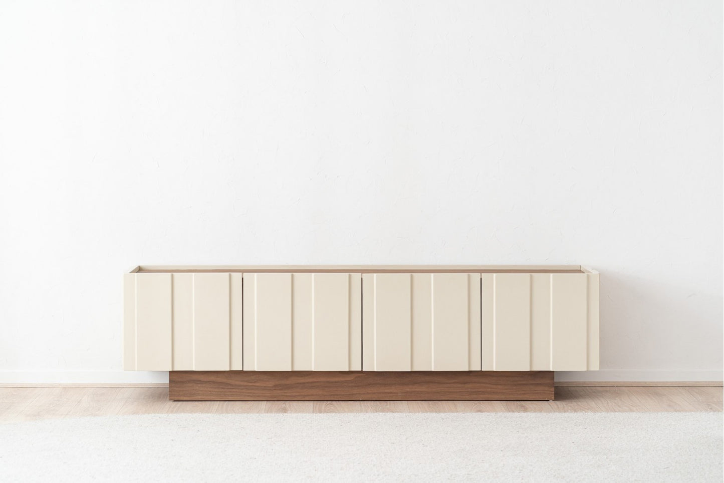 Un mueble TV de madera de nogal lacado beige brillo OBI (198×45×53 cm), diseño moderno, con paneles verticales y base nogal, se apoya en una pared blanca sobre suelo de madera clara.