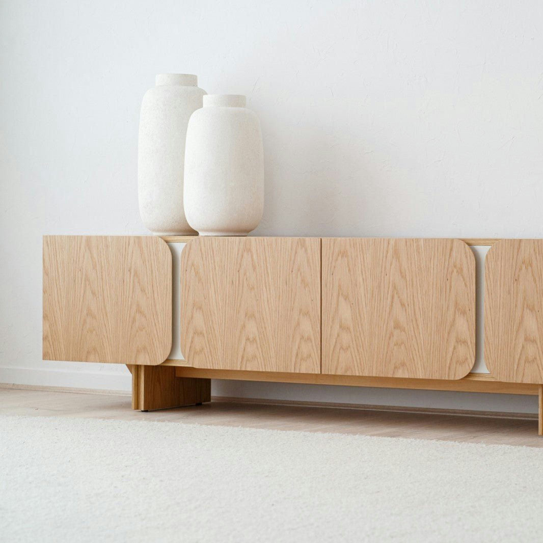 El Mueble TV en DM y roble natural detalles gris arena Curva, un aparador minimalista de madera con un tono arena mate, sostiene dos grandes jarrones de cerámica blanca. Colocado contra una pared blanca lisa y un piso alfombrado claro, irradia serenidad y sofisticación.