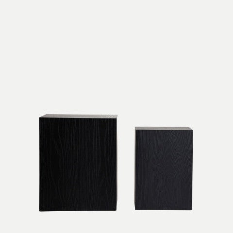 Dos cajas de madera negras de la Mesa auxiliar cuadrada en madera de abeto Peana Amilly (2 Piezas), elaboradas con elegante madera de abeto, se colocan una al lado de la otra sobre un fondo blanco, mostrando su estilo minimalista.