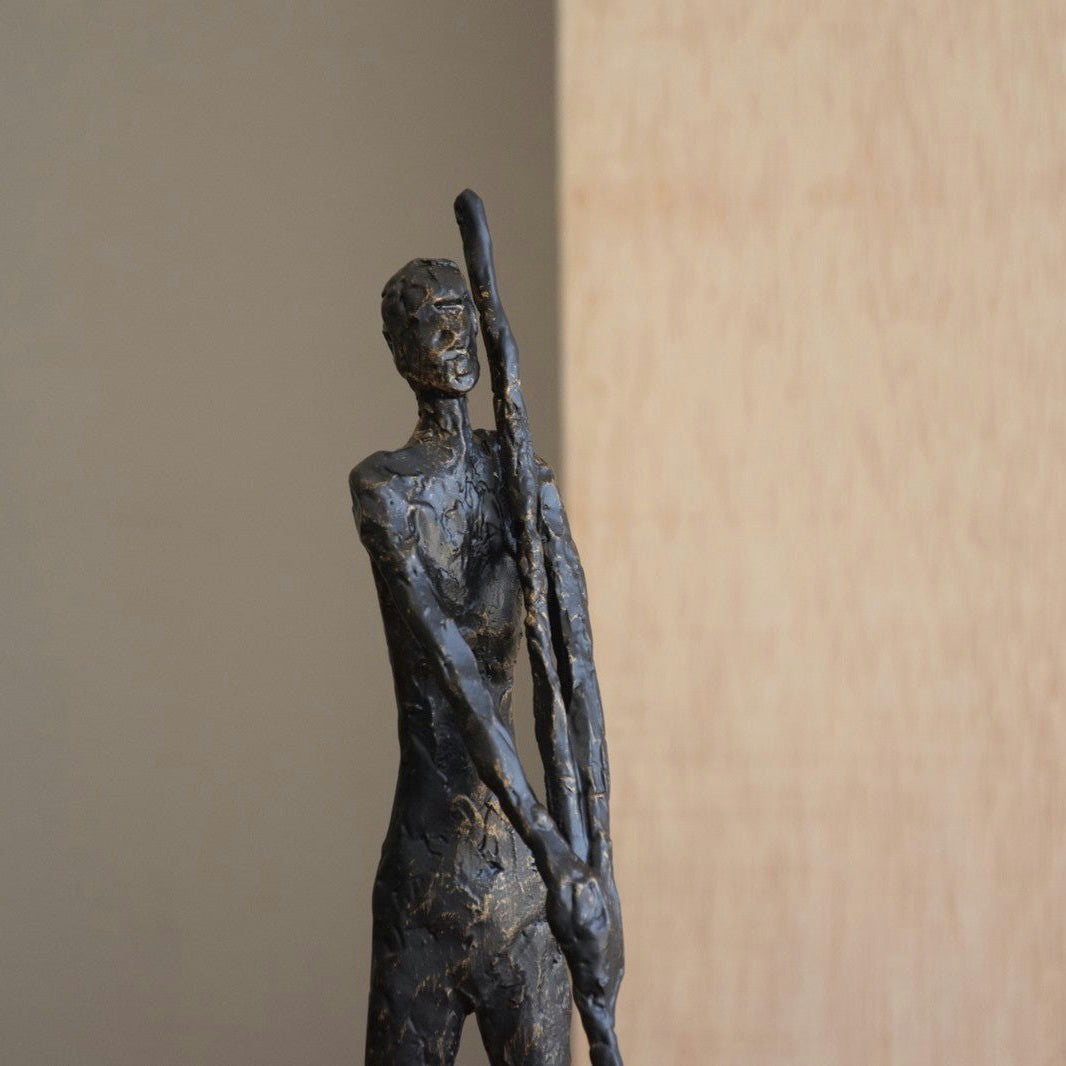 La Figura decorativa de metal en bronce envejecido Hombre N3 muestra una escultura abstracta de un humano esbelto con un poste, sobre fondo neutro, perfecta para la decoración artística.